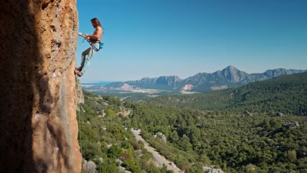 Seitenansicht eines starken attraktiven Bergsteigers, der sich bereit macht, von einer senkrechten Klippe am oberen Seil herabzusteigen. felsigen Bergkamm und klaren blauen Himmel auf dem Hintergrund. gesunder Lebensstil und sportliche Betätigung — Stockvideo