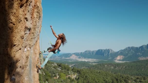 Vahva ja istuva mies kalliokiipeilijä roikkuu köydellä pystysuoralla kalliolla, katsoo ylös ja näyttää käsin. kaunis maisema kivinen vuorijono ja kirkas sininen taivas taustalla — kuvapankkivideo