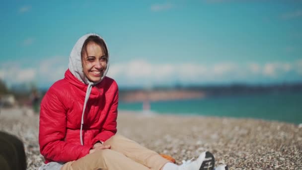 Young woman in red jacket and hoodie sitting on pebble sea beach at windy sunny day and picking up pebble stones — Stock Video