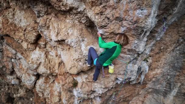 Slow motion aerial view strong female rock climber climbs on overhanging cliff by hard route with rock tufas. woman makes tough effort and reaches holds for leg. — Stock Video