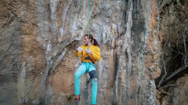 Slow motion van vrolijke knappe man rock klimmer in gele sweater met lang haar opknoping op touw en geven high five na succesvolle beklimming van de route op klif. — Stockvideo