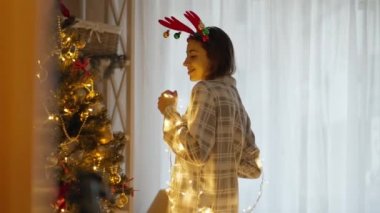 Mutlu, güzel bir kadının portresi pijama giyiyor ve geyik boynuzları takıyor Noel ışıklarıyla Noel ağacıyla evde dans ediyor. xmas ve mutlu yıllar konsepti