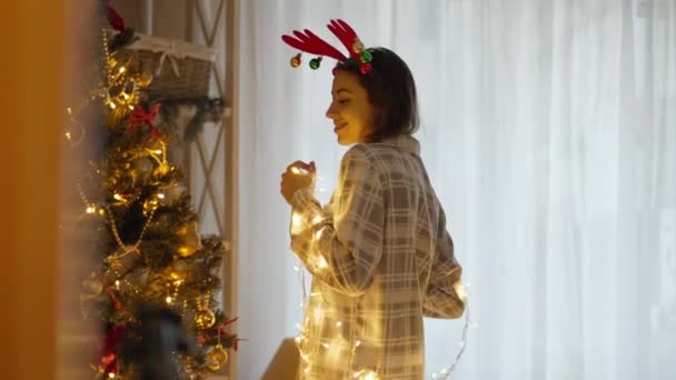 Ritratto di felice allegra bella donna che indossa pigiama e corna di renna fascia ballare con luci di Natale a casa accogliente con albero di Natale. Natale e felice anno nuovo concetto — Video Stock