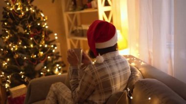 Pijamalı ve kırmızı Noel Baba şapkalı kadın koltukta oturuyor ve Noel arifesinde ve yeni yılda rahatlamak için fincandan sıcak çikolata içiyor. Rahat bir evde ışıklar ve yılbaşı ağacıyla süslenmiş bir kız.