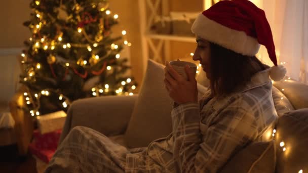 Жінка в піжамі і червоному капелюсі Санти сидить на дивані і п'є гарячий шоколад з чашки, відпочиваючи в Святвечір і Новий рік. дівчина у затишному оздобленому домі з сяйвом та кришталевими деревами. — стокове відео
