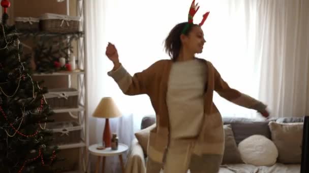 Young cheerful pretty woman wearing reindeer antlers headband dancing with Christmas lights at cozy home — Stock Video