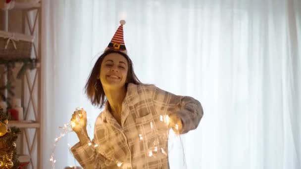 Retrato en cámara lenta de 4k de la alegre mujer bonita y feliz usando pijama y la pequeña diadema Elf Cap bailando con luces de Navidad en la acogedora casa con árbol de Navidad. Navidad y feliz año nuevo concepto — Vídeo de stock