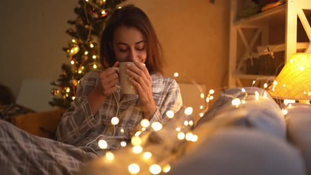 Kamera zoomning till glad drömmande flicka i pyjamas sitter på soffan med ljus, dricka varm choklad i varma festliga rum. kvinna på mysiga inredda hem med ljus och julgran på julafton — Stockvideo