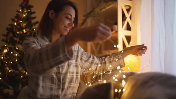 Портрет счастливой мечтательницы, украшающей диван огнями на Рождество. женщина в пижаме на уютном украшенном доме с огнями цепь и елка на заднем плане — стоковое видео