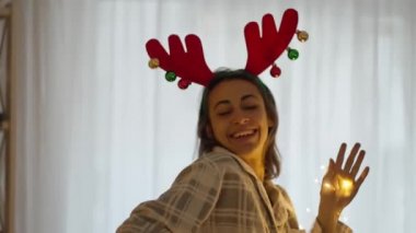 Ren geyiği boynuzu takan neşeli, güzel bir kadın evinde Noel ışıklarıyla dans ediyor. xmas ve mutlu yıllar konsepti