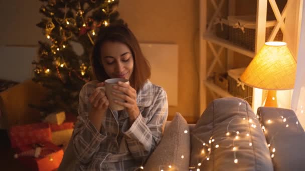 Kamera zoomning till glad drömmande flicka i pyjamas sitter på soffan med ljus, dricka varm choklad i varma festliga rum. kvinna på mysiga inredda hem med ljus och julgran på julafton — Stockvideo
