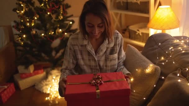 Повільний рух щаслива насолоджена красива жінка відкриває різдвяну подарункову коробку на Різдвяному святкуванні вдома. Здивована дівчина в милій піжамі шукає подарункову коробку зі світлом всередині — стокове відео