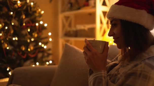 Donna in pigiama e cappello da Babbo Natale rosso seduta sul divano e che beve cioccolata calda dalla tazza, rilassarsi la vigilia di Natale e Capodanno. ragazza in accogliente casa decorata con luci e albero di Natale — Video Stock