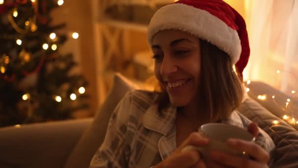 Kvinna i pyjamas och röd tomte hatt sitter på soffan, titta på TV och dricka varm choklad från mugg, koppla av på julafton, nyår. flicka på mysiga inredda hem med ljus och julgran — Stockvideo