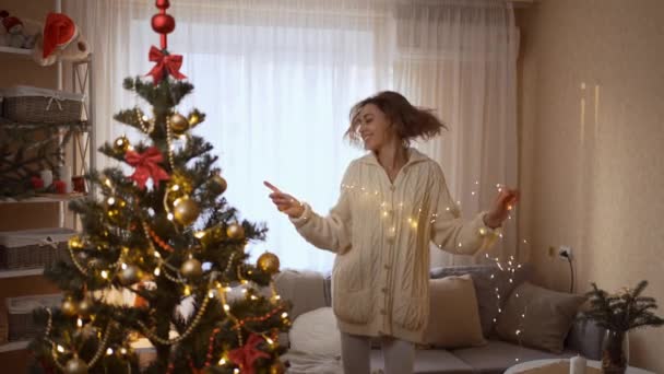 4k slow motion of happy pretty woman dancing with Christmas lights at cozy festive home with christmas tree. xmas and happy new year at home athmosphere concept — Stock Video