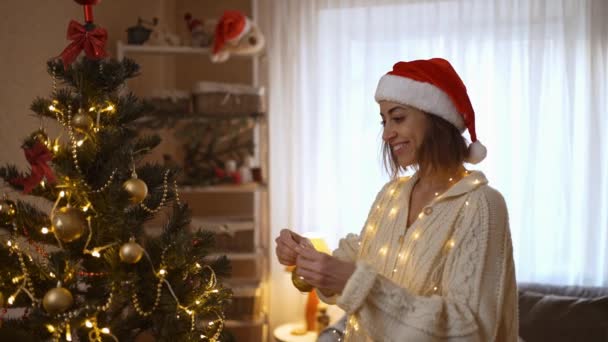 Ritratto al rallentatore p di donna felice a Santa Hat appende palla d'oro sul ramo dell'albero di Natale. decorazione della casa e la preparazione per festeggiare il Natale e Capodanno a casa — Video Stock