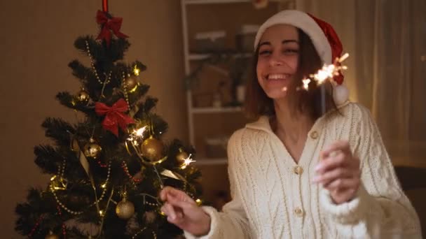 4k Zeitlupe glücklich lächelnde junge Frau mit Weihnachtsmütze tanzt mit Wunderkerze zu Weihnachten oder Neujahr im gemütlichen Haus — Stockvideo