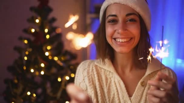 Close up πορτρέτο ευτυχισμένη χαρούμενη νεαρή γυναίκα σε Σάντα καπέλο με sparklers την παραμονή των Χριστουγέννων ή την Πρωτοχρονιά στο άνετο σπίτι με χριστουγεννιάτικο δέντρο. κινηματογραφική 4k αργή κίνηση — Αρχείο Βίντεο