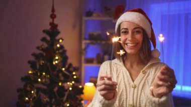 Noel arifesinde ya da Noel ağacıyla dolu sıcak bir evde yanan maytapları olan mutlu yüzlü, Noel Baba şapkalı kadın. Sinematik 4k ağır çekim