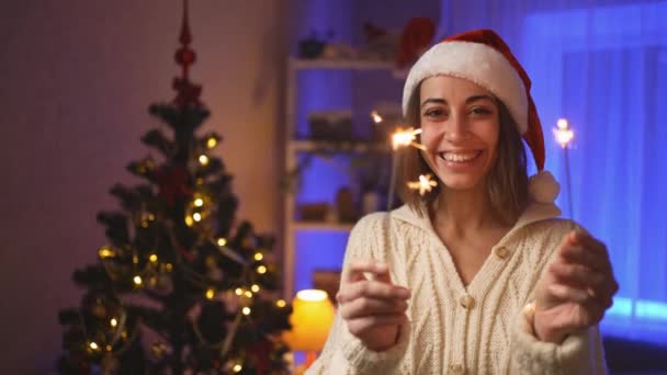 Cerrar la cara feliz sonriente mujer en el sombrero de Santa con bengalas ardientes en Nochebuena o Año Nuevo en la casa acogedora con árbol de Navidad. Cinemática 4k cámara lenta — Vídeos de Stock