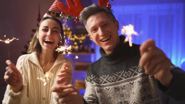 Щаслива пара, жінка у святковій капці Ельфа і чоловік у червоних оленячих рогах, дивлячись на камеру з блискітками на Різдво або Новий рік у затишному будинку. 4k повільний рух — стокове відео
