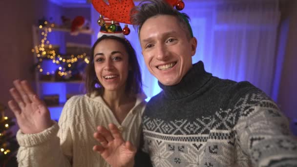 POV primeira pessoa ver feliz alegre casal animado, mulher em festivo Elf Cap e homem de renas vermelhas chifres headband tomando selfie no fundo da casa decorada com árvore de natal — Vídeo de Stock