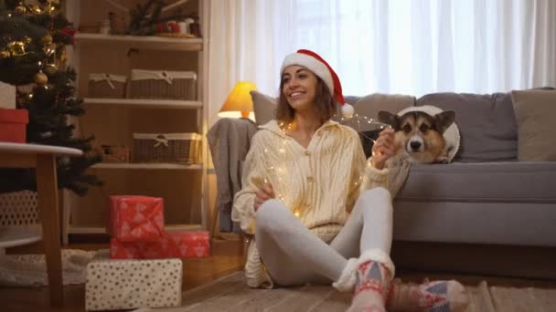 Emotiva joven sonriente en Santa sombrero sosteniendo luces, y su divertido perro Corgi se sienta en el sofá en la casa decorada festiva con árbol de Navidad. 4k cámara lenta b-roll material de archivo concepto de Año Nuevo — Vídeos de Stock