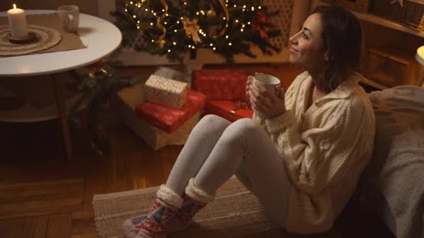 Slowmotion porträtt drömmande flicka i stickad tröja sitter på golvet på soffan, dricka varm choklad i varma festliga rum. kvinna på mysiga inredda hem med ljus och julgran på julafton — Stockvideo