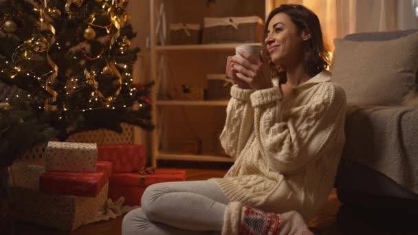 B-roll cámara lenta bonita mujer usando cálido lindo invierno blanco suéter de punto beber chocolate caliente y relajarse sentado en el suelo en la casa decorada con luces y árbol de Navidad — Vídeos de Stock
