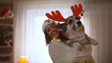 Noel Baba şapkalı neşeli kadının 4k yavaş çekimde elinde komik Galli Corgi köpeğinin boynuzları ve rahat festival evinde boynuzları var. Hayvan konseptiyle evde tatiller