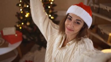 POV, Noel Baba Şapkalı mutlu ve heyecanlı kadını, dekore edilmiş evin arka planında Noel ağacıyla selfie çekerken görüyor.