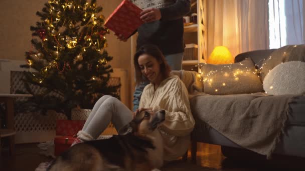 Familj firar jul med söt Corgi hund på festliga inredda mysiga hus. glad leende kvinna stroking hund och få närvarande i presentförpackning från kärleksfull man på nyårsfirande hemma — Stockvideo