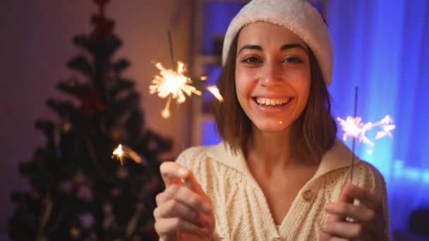 B-roll close up πορτρέτο χαμογελώντας χαρούμενη κοπέλα σε Σάντα καπέλο με καύση sparklers την παραμονή των Χριστουγέννων ή την Πρωτοχρονιά στο άνετο σπίτι με χριστουγεννιάτικο δέντρο. κινηματογραφική 4k αργή κίνηση — Αρχείο Βίντεο