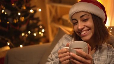 Pijamalı ve kırmızı Noel Baba şapkalı bir kadın kanepeye oturmuş televizyon izliyor ve kupasından sıcak çikolata içiyor. Noel arifesinde, Noel arifesinde dinleniyor. Rahat bir evde ışıklar ve yılbaşı ağacıyla süslenmiş bir kız.