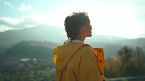 Радостная девушка свободы в желтой куртке стоит с распростертыми объятиями вокруг гор и холмов, солнце светит над рукой, чувствуя себя счастливым — стоковое видео