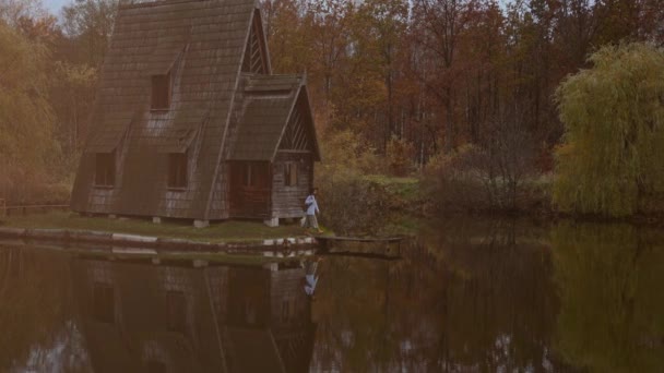 Panoramique b-roll de paysage panoramique d'automne. Maison en bois au bord du lac au milieu de la forêt, jolie fille marchant sur une jetée en bois. réflexion foliaire dans l'eau — Video