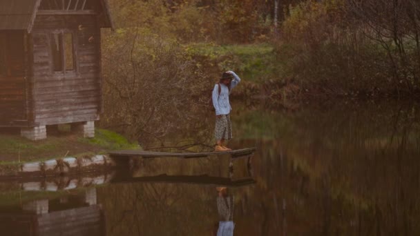 Vecchia casa in legno capanna sul lago tra i boschi autunnali, donna passeggiante vicino al molo di legno. Riflessione fogliare in acqua — Video Stock