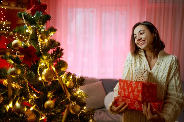 Brunette nainen vastaanottaa lahjoja jouluna, seisoo koristeltu joulukuusi olohuoneessa — kuvapankkivalokuva