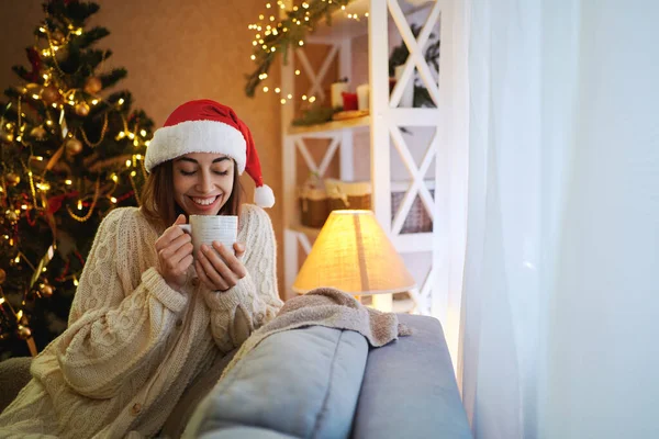 Muotokuva Joulupukki tyttö nauttia kuppi teetä istuessaan sohvalla sisustettu olohuone — kuvapankkivalokuva
