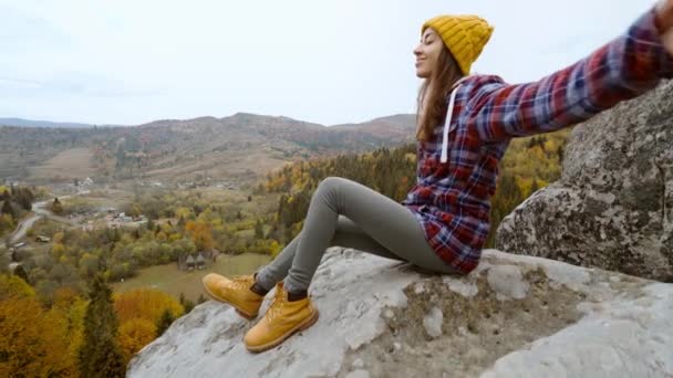 POV první osoba pohled na ženu turistka v teplé ležérní oblečení sedí na okraji útesu na pozadí lesa s domy pod hřebenem hory a zvedá ruce — Stock video
