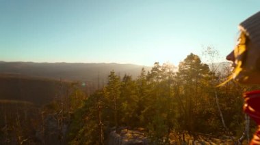 Kırmızı ceketli ve bereli kendine güvenen genç bir kadın. Kız el kaldırıyor, doğanın tadını çıkarıyor ve gün batımında dağın tepesinde yaptığı yürüyüş ile kendinden emin ve mutlu görünüyor.