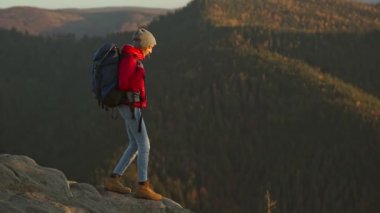 Yavaş çekim. Kırmızı yalıtılmış ceket ve kot pantolon giyen maceraperest kadın yürüyüşçü gün batımında ormanda kayalık bir dağdan aşağı iniyor.