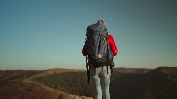 Cámara lenta vista trasera Joven aventurera mujer excursionista mochilero en una chaqueta aislante rojo y jeans trekking por una montaña rocosa al atardecer por encima del bosque. womn raisinh manos en la cima de acantilado montaña — Vídeo de stock