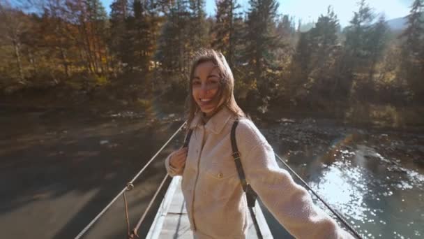 Slow motion portrait of happy beautiful young woman tourist standing on wooden Suspension Bridge hanged over mountain river. amazing landscape with spruces, mountains and sky on background — Stock Video