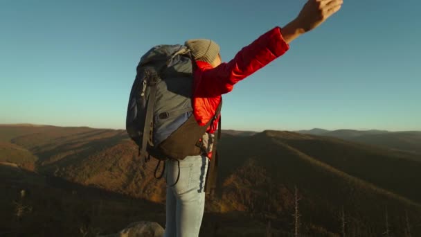 Zpět pohled šťastná žena turistka batohem na sobě červené sako a pěší turistika zvedá ruce do nebe. dívka těší pohled na hory a lesy na vrcholu hory při podzimním západu slunce — Stock video