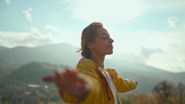 Jovem mulher vestindo uma jaqueta amarela levantando os braços para o céu azul e desfrutando da vista acima das nuvens no topo de uma montanha — Vídeo de Stock