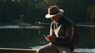30 'ların Hiker fotoğrafçısı hippi adam ormanda Selfie Lake View çekiyor. Çağdaş Seyahat Videografı Şapka ve Sırt Çantası Doğada Duruyor ve Akıllı Telefon Kamerasında Kendini Çekiyor