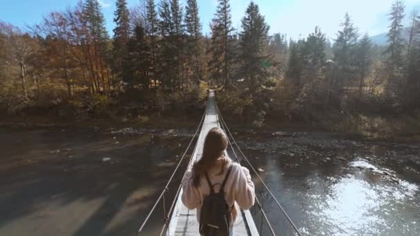 スローモーションカメラは、マウンテン川に架かっている木製の吊り橋を歩くヴィンテージジャケットとジーンズの女性観光客に続きます。背景にはスプルースや山や空を背景にした素晴らしい風景 — ストック動画