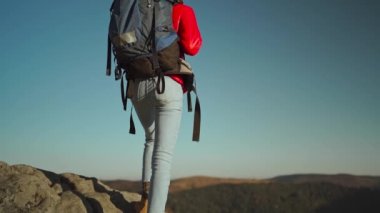 Yavaş çekim dikiz aynası. Kırmızı yalıtılmış ceket ve kot pantolon giymiş maceraperest kadın yürüyüşçü. Ormanın yukarısındaki kayalık bir dağda gün batımında yürüyüş yapıyor. Uçurumun tepesinde ellerinizi kaldırın.
