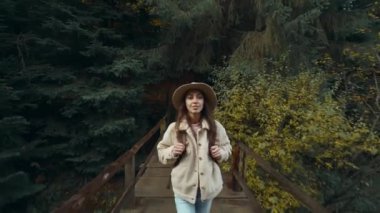Gündelik kıyafetleri ve şapkası olan çekici hippi kadın sonbaharda ormanda yürüyüş yapıyor. Turist bir gezgin ormanda yürüyüş yapıyor. Dişi gezgin dağ gölündeki eski ahşap köprüde yürüyor.